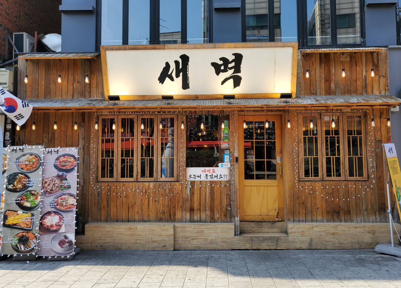 낮에 본 새벽 외관 — 목조 건물 앞에 메뉴 사진 입간판과 태극기가 서 있고, 유리문 너머로 주황색 내부가 비친다