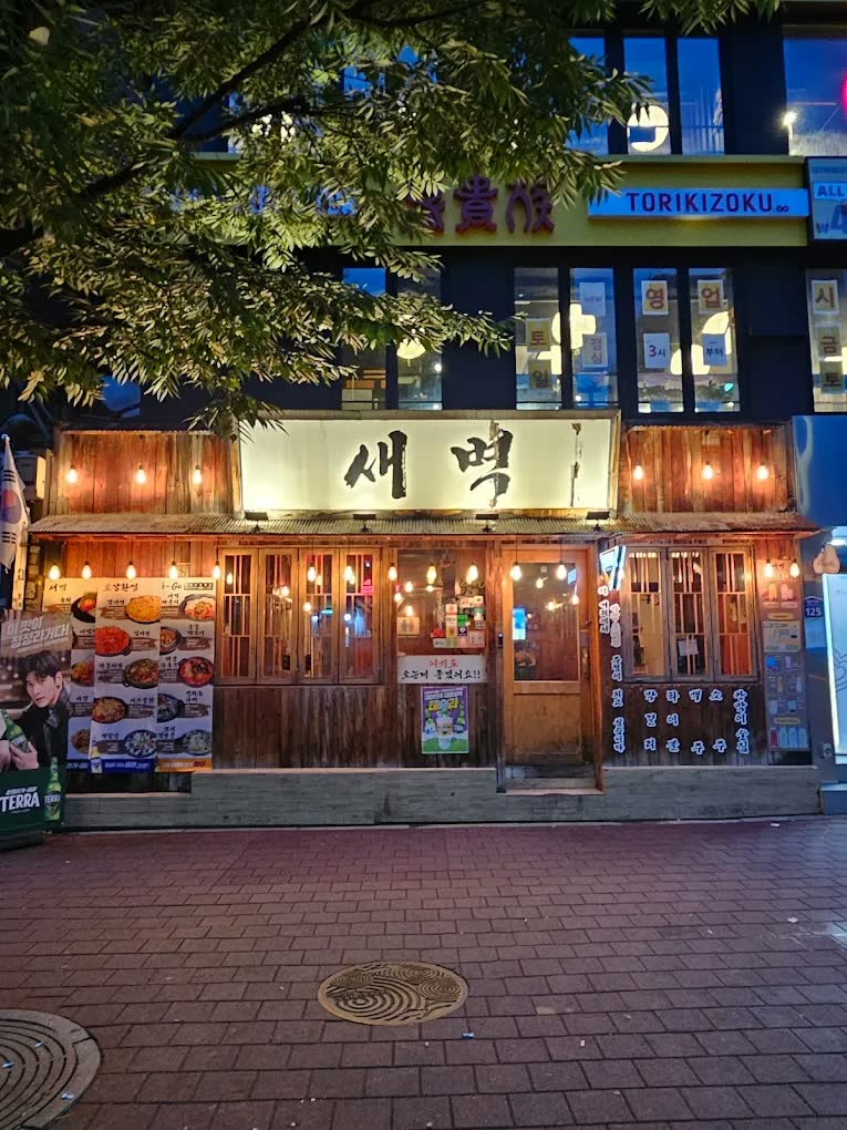 밤의 어울마당로에서 본 새벽 외관 — 목조 파사드 위에 붓글씨로 쓴 ‘새벽’ 간판이 조명을 받고, 처마 아래로 전구가 줄지어 걸려 있다